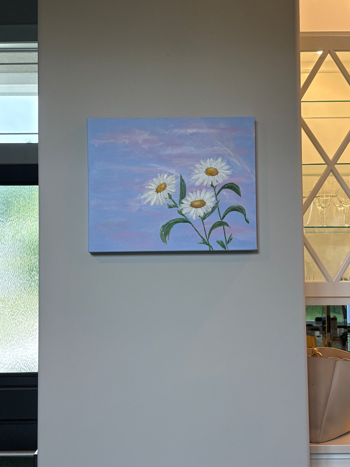 rectangle canvas painting of three white daisies with yellow centers and green stems against a soft blue and pink sky, displayed on a light gray wall in an interior setting.