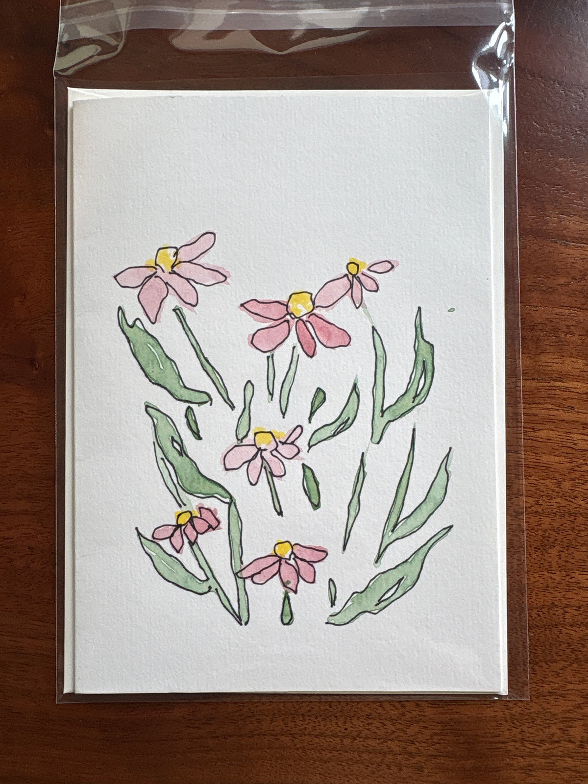 A square white art print or card featuring a hand-drawn floral illustration of pink flowers with yellow centers and green leaves, sealed in a clear plastic sleeve and placed on a dark wooden surface.