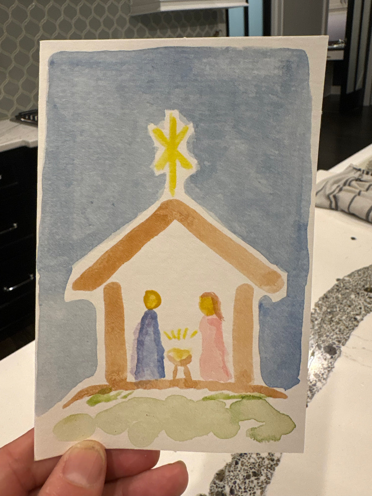 A hand holding a small watercolor painting of a nativity scene with a stable, blue sky, and a yellow star, photographed in a real indoor environment.