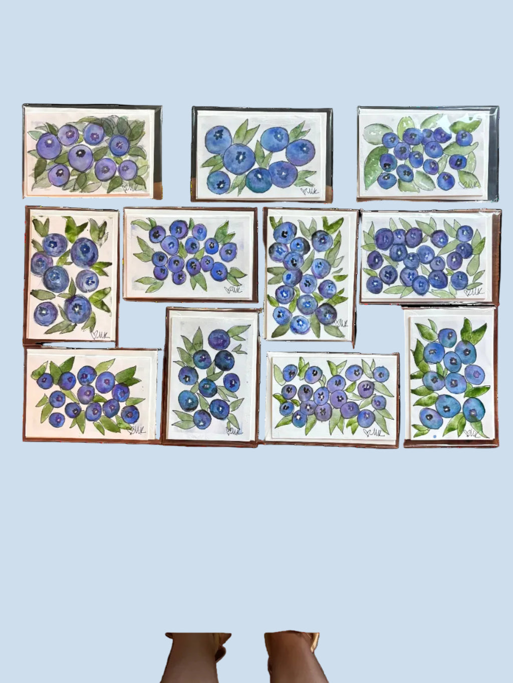 A set of nine small square watercolor paintings of blueberries arranged on a wooden table. The paintings feature various styles and details of blueberry clusters in shades of blue, purple, and green on white backgrounds.
