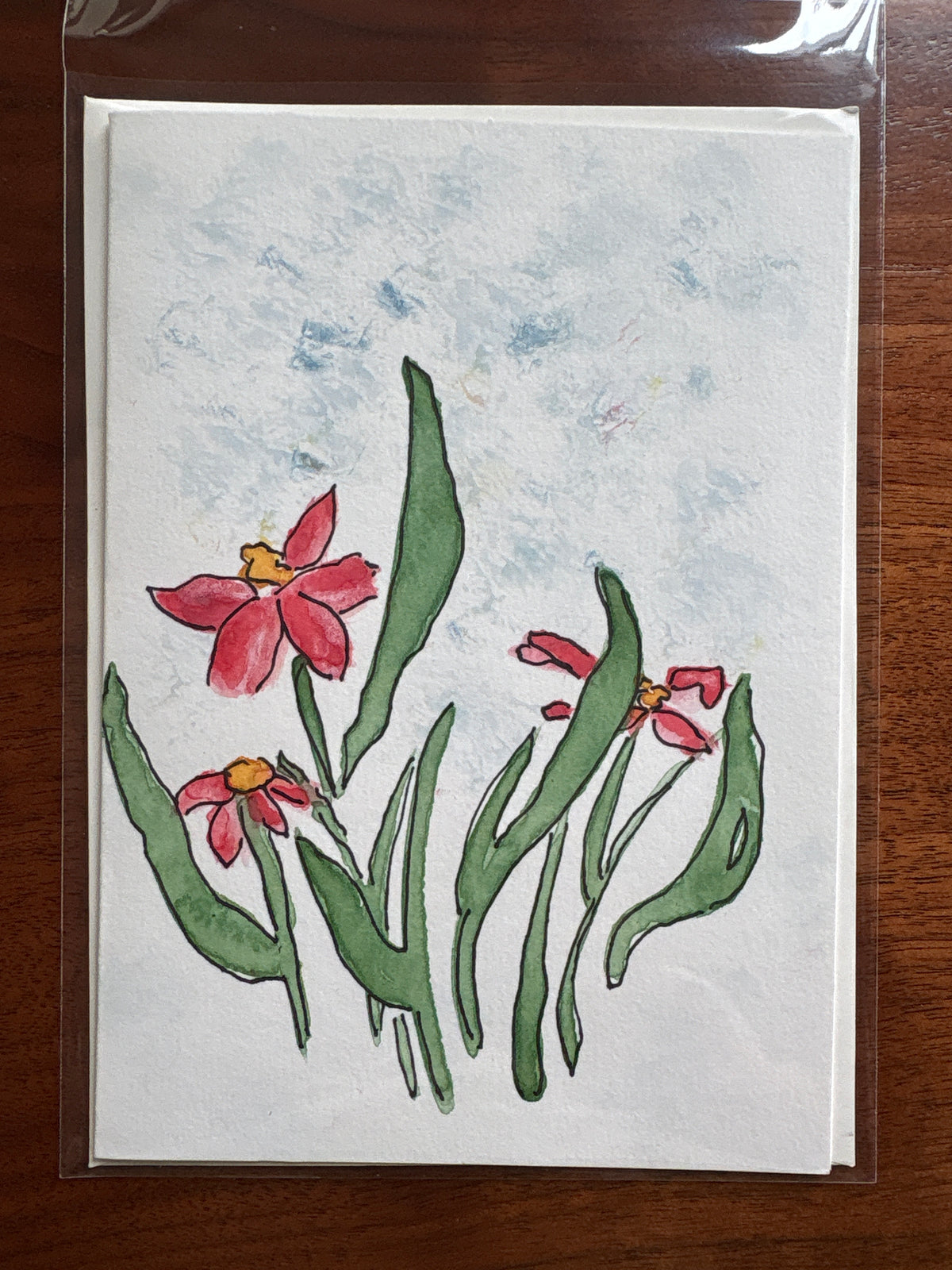 A square watercolor-style floral art print or card featuring pink-red flowers with green leaves on a white textured background, photographed inside a clear plastic sleeve on a brown wooden surface.