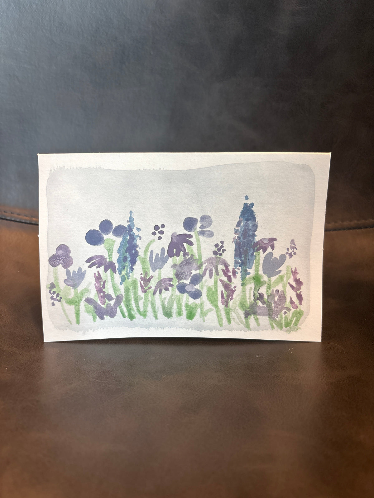 A small square watercolor painting of purple and blue flowers with green stems on a white paper background, displayed on a dark brown leather surface against a dark gray wall.